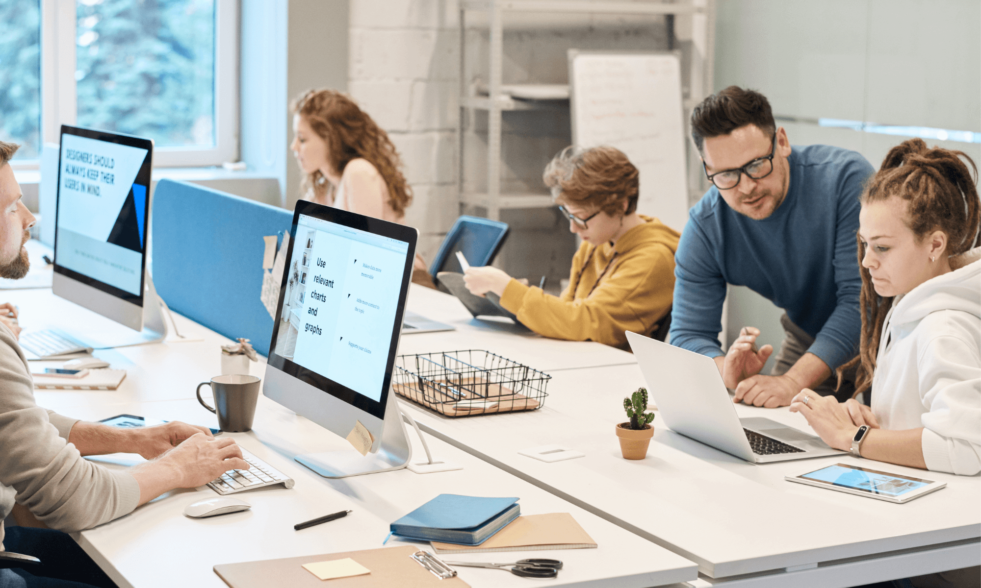 A man is helping a female apprentice understand something on a laptop. They are working around a large communal desk in an office